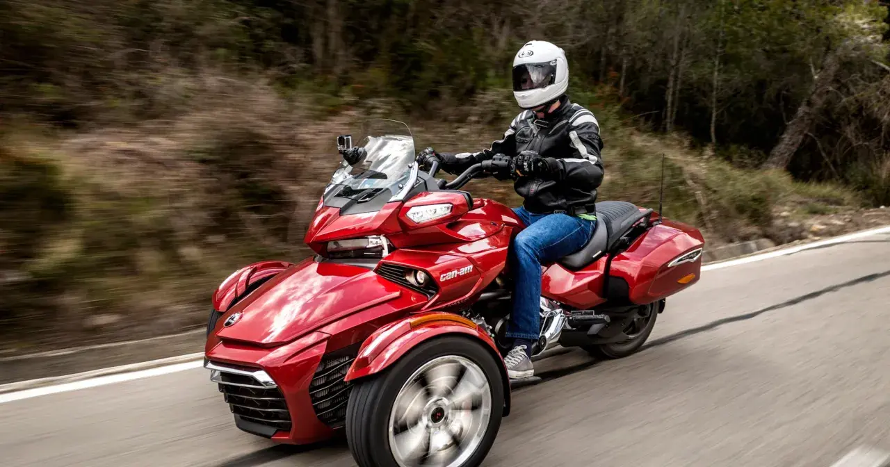 Czerwony pojazd trójkołowy z kierowcą w kasku i kurtce, jadący po krętej drodze. Idealny dla tych, którzy szukają wrażeń, ale nie mają prawa jazdy na motocykl.
