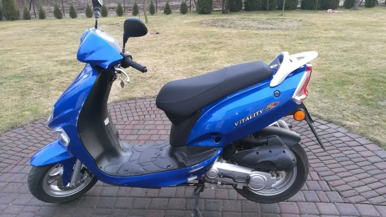 Błękitny skuter Kymco Vitality 50 2T stoi na brukowanym podjeździe, gotowy do jazdy.