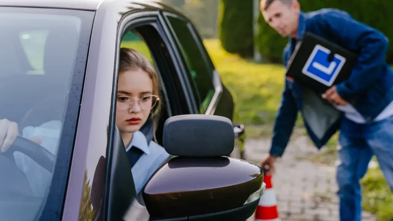 Dziewczyna za kierownicą auta szkoleniowego, instruktor z tablicą "L" obok. Spełnienie wymagań na prawo jazdy kat. A.