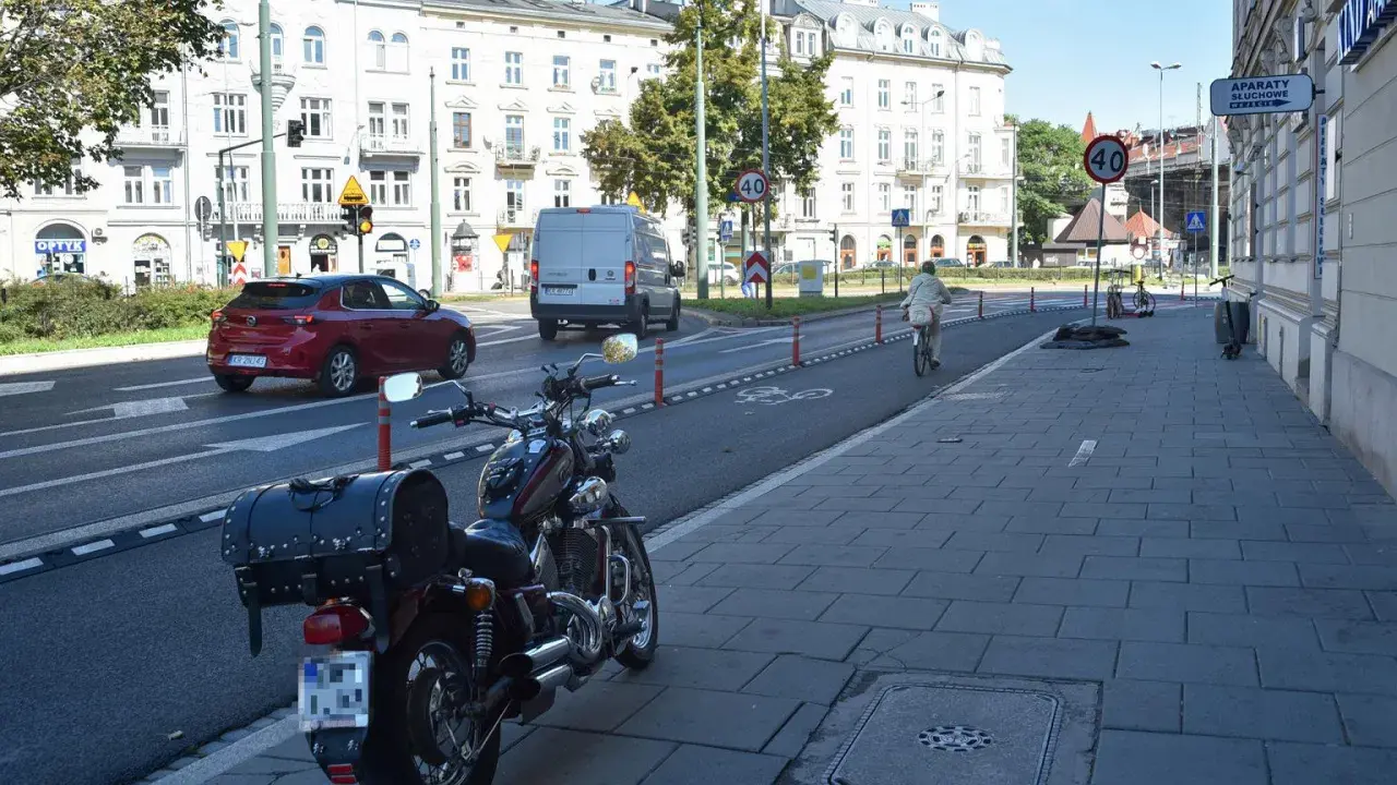 Motocykl zaparkowany na chodniku, obok drogi z wyznaczonym pasem rowerowym. W tle budynki i ruch uliczny, w tym czerwony samochód i biały van.
