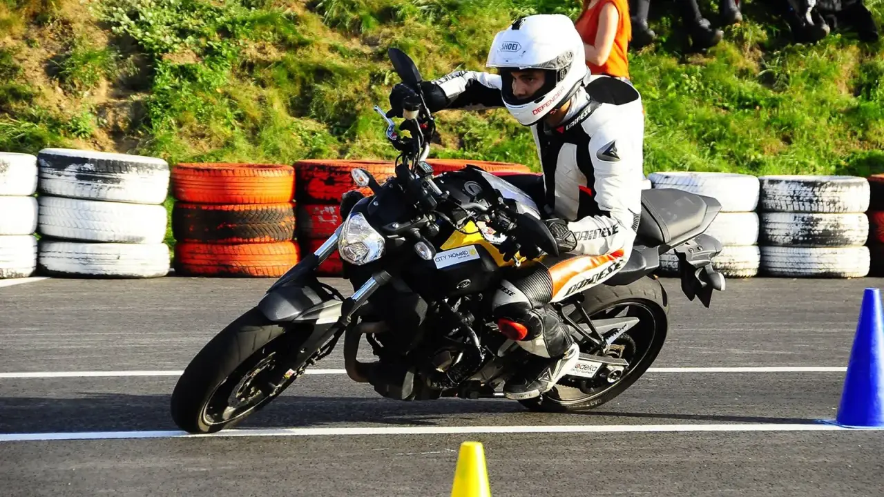Motocyklista w kombinezonie i kasku wykonuje zakręt, przyjmując sportową pozycję na motocyklu.
