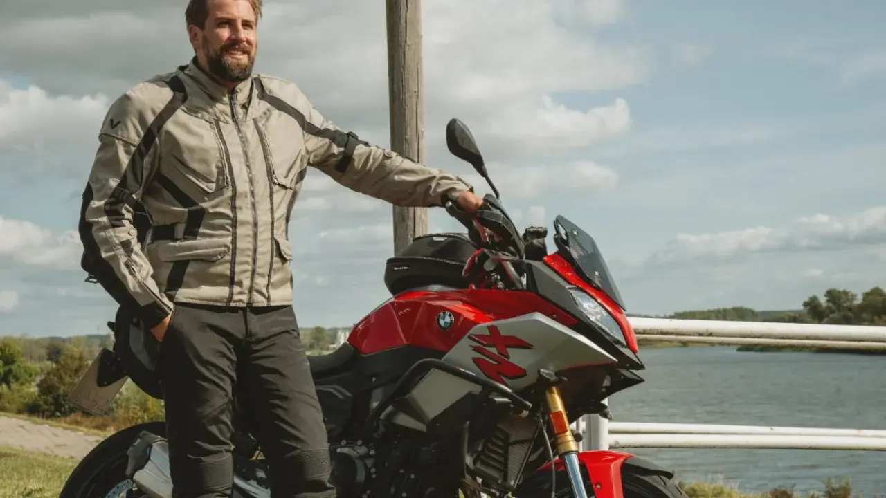 Uśmiechnięty motocyklista w kurtce od polskiego producenta odzieży motocyklowej, oparty o czerwoną maszynę nad jeziorem.