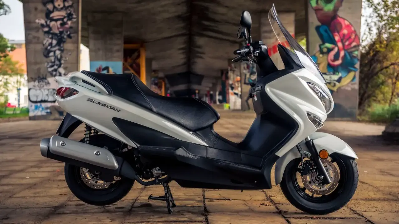 Biały skuter Suzuki Burgman 125, z czarnym siedzeniem, zaparkowany pod mostem z graffiti. Poznaj suzuki burgman 125 dane techniczne.