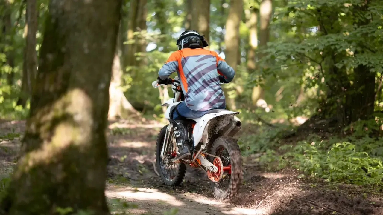 Motocyklista w kasku i pomarańczowej bluzie pędzi leśnym szlakiem. Od ilu lat można jeździć crossem? Odpowiedź zależy od przepisów i umiejętności.