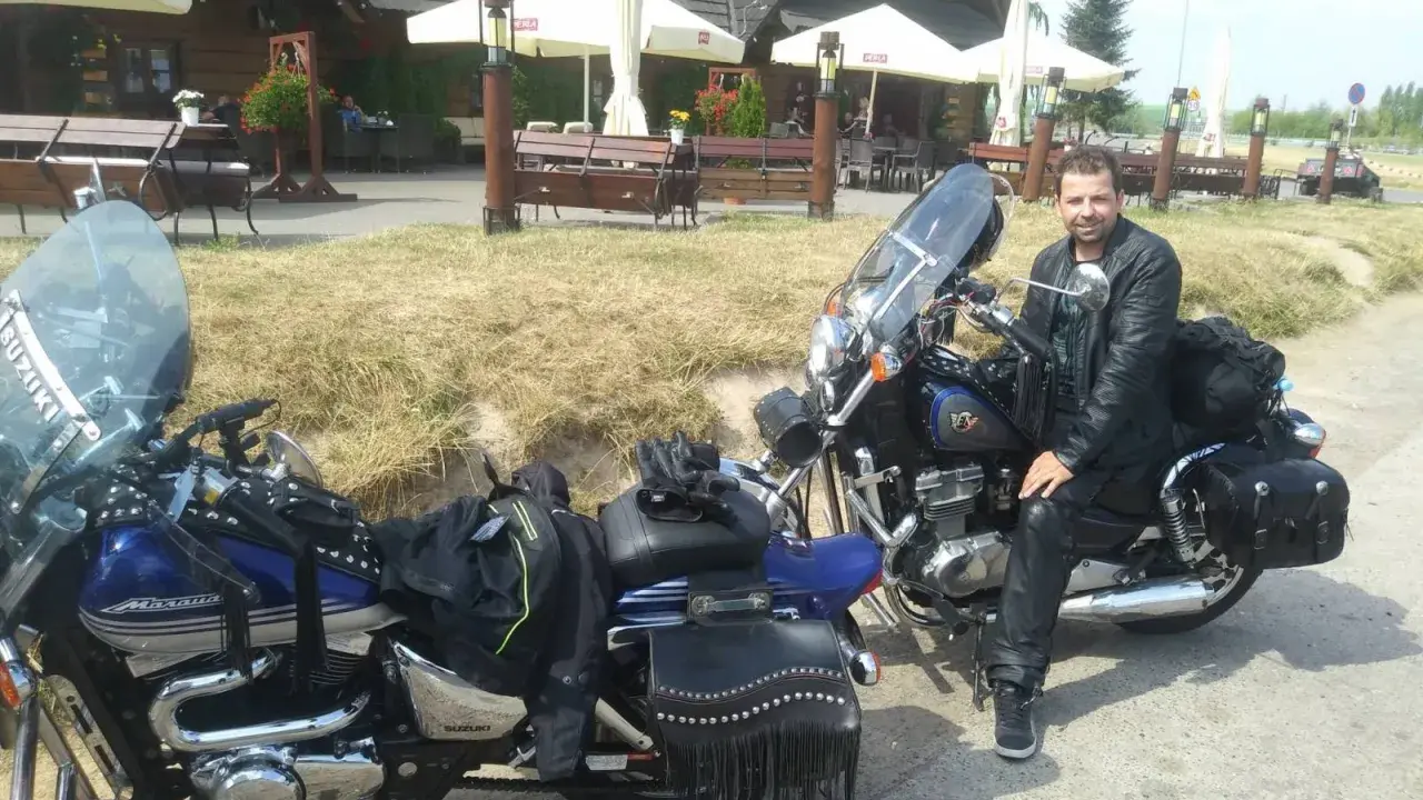 Motocyklista w skórzanej kurtce i spodniach odpoczywa przy swoim motocyklu. W tle karczma "Bida", idealne miejsce na postój podczas odkrywania **tras motocyklowych lubelskich**.