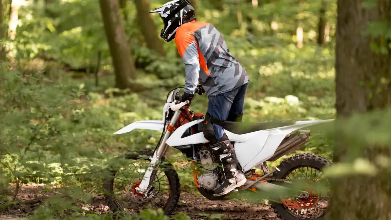 Motocyklista w kasku i pomarańczowej bluzie jedzie na białym crossie przez leśną ścieżkę.