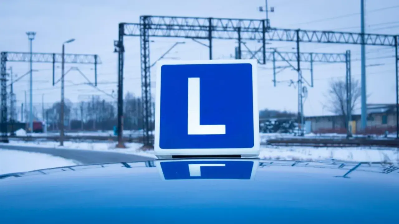 Tablica z literą "L" na dachu samochodu, symbolizująca naukę jazdy i teorię na prawo jazdy. W tle zimowy krajobraz z torami kolejowymi.