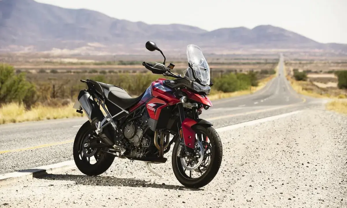 Czerwony motocykl Triumph Tiger 900 GT Pro zaparkowany na poboczu pustej drogi w krajobrazie z górami w tle.