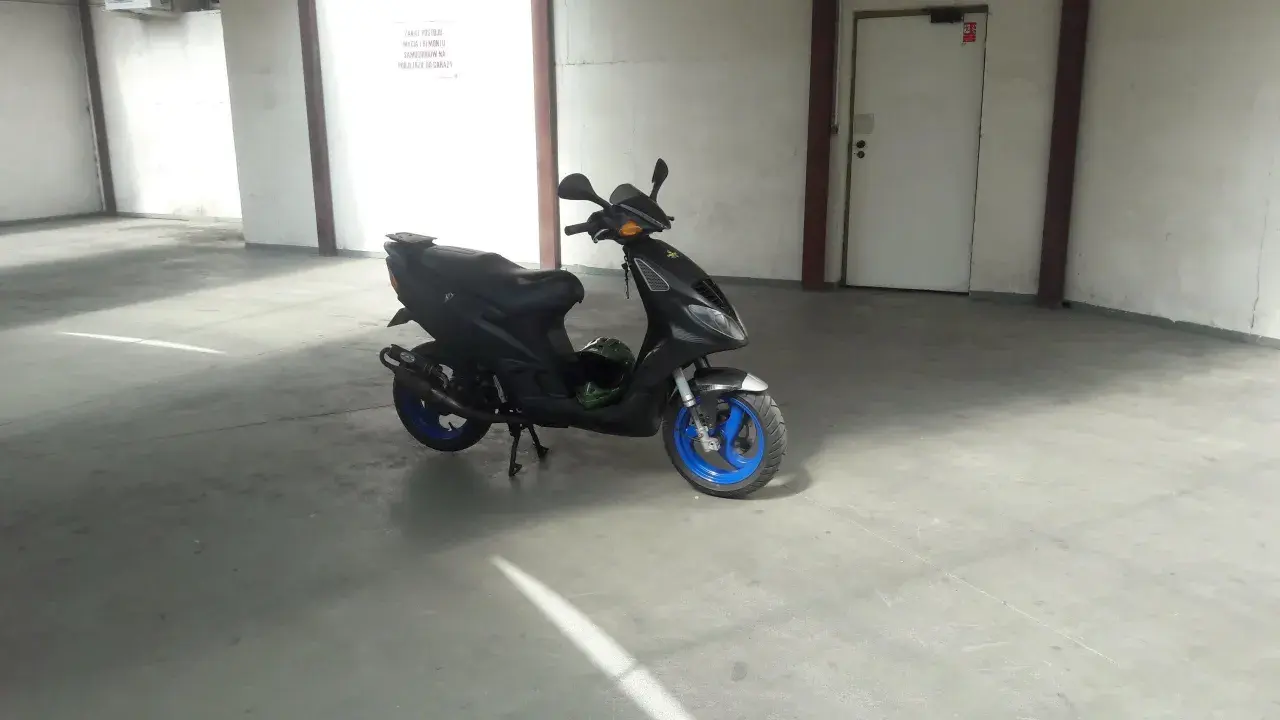 Czarny skuter Piaggio NRG MC3 z niebieskimi felgami stoi na betonowej posadzce w garażu. Obok niego leży zielony kask.