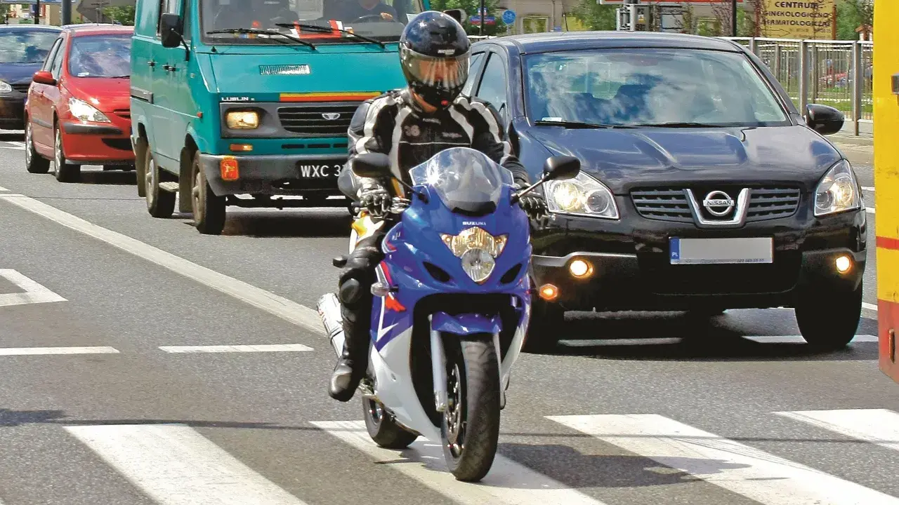 Motocyklista w kasku i kombinezonie na pasach. Obok samochody i furgonetka. Pamiętaj o przeglądzie, bo jaka kara za brak przeglądu może być dotkliwa.