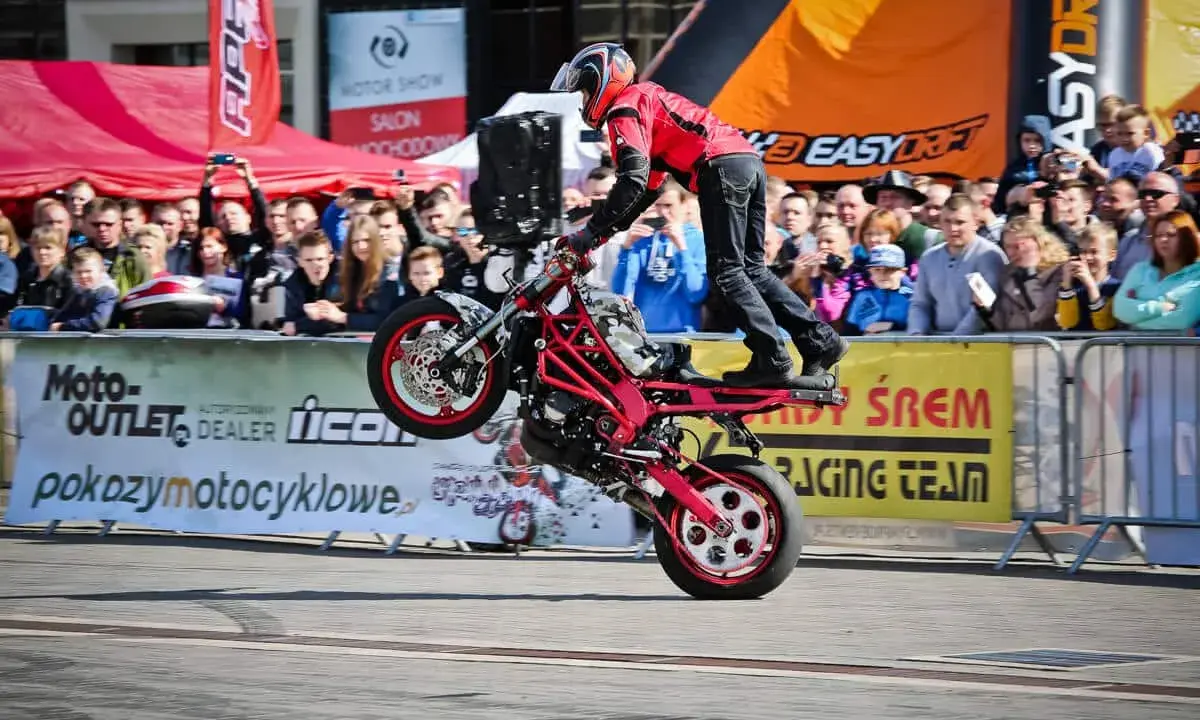 Motocykl do stuntu na tylnym kole, kierowca w czerwonej kurtce i kasku, tłum widzów na tle banerów.