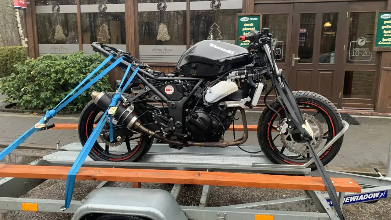 Czarny motocykl Kawasaki na przyczepce, zabezpieczony pasami. W tle budynek z napisem "Leśny Staw". Przepisy dotyczące przyczepki do motocykla są kluczowe dla bezpiecznego transportu.