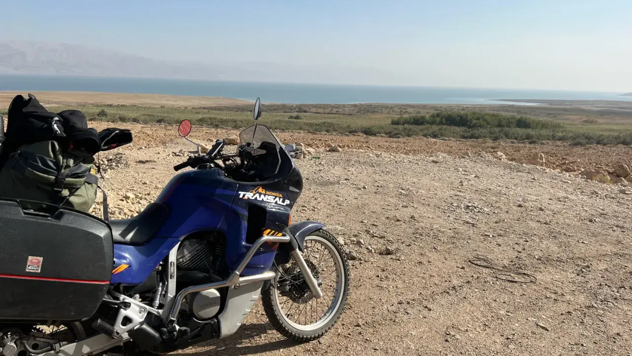 Motocykl Honda Transalp z bagażami gotowy do podróży, z widokiem na Morze Martwe. Dokumenty, jak **carnet de passage**, czekają na przygodę.
