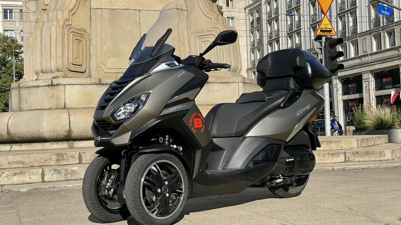 Trójkołowy skuter Peugeot Metropolis 400 na tle zabytkowej kamienicy i pomnika.