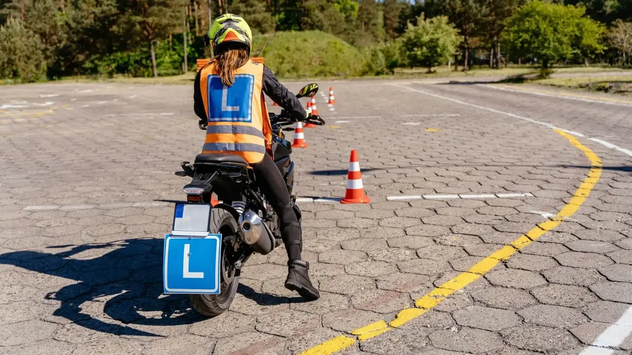 Nauka jazdy motocyklem na placu manewrowym. Osoba w kamizelce odblaskowej z literą "L" na motocyklu.