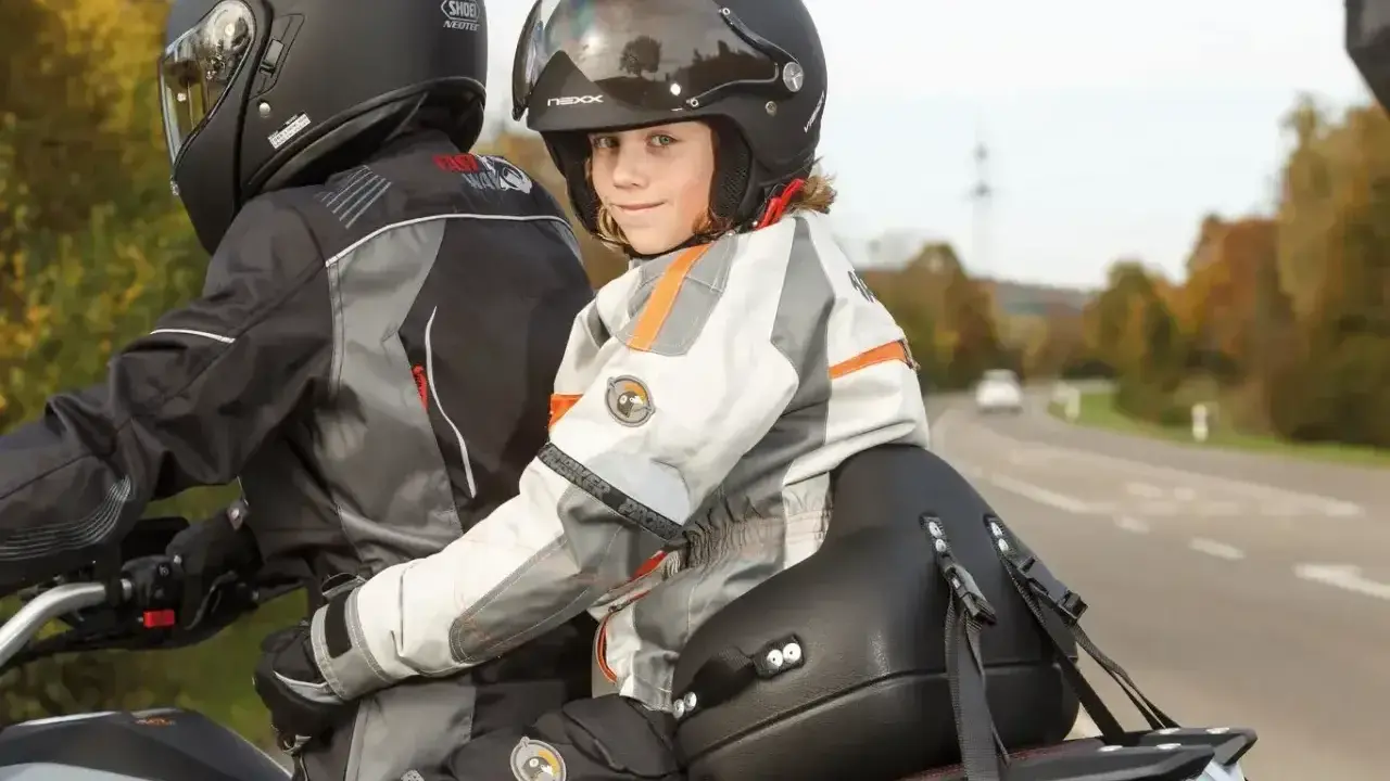 Uśmiechnięte dziecko na motocyklu, w kasku i kurtce, siedzi za kierowcą.