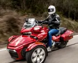 Czerwony pojazd trójkołowy z kierowcą w kasku i kurtce, jadący po krętej drodze. Idealny dla tych, którzy szukają wrażeń, ale nie mają prawa jazdy na motocykl.