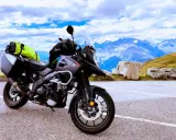 Szary motocykl Suzuki V-Strom, idealny motocykl do turystyki we dwoje, zaparkowany na tle majestatycznych gór i błękitnego nieba.