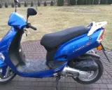 Błękitny skuter Kymco Vitality 50 2T stoi na brukowanym podjeździe, gotowy do jazdy.