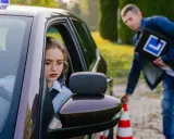 Dziewczyna za kierownicą auta szkoleniowego, instruktor z tablicą "L" obok. Spełnienie wymagań na prawo jazdy kat. A.