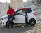 Młoda para przy białym samochodzie z czerwonym dachem. Ona opiera się o maskę, on siedzi za kierownicą. Idealni kandydaci na am1 prawo jazdy.