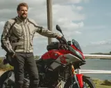 Uśmiechnięty motocyklista w kurtce od polskiego producenta odzieży motocyklowej, oparty o czerwoną maszynę nad jeziorem.