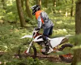 Motocyklista w kasku i pomarańczowej bluzie jedzie na białym crossie przez leśną ścieżkę.
