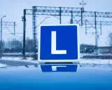 Tablica z literą "L" na dachu samochodu, symbolizująca naukę jazdy i teorię na prawo jazdy. W tle zimowy krajobraz z torami kolejowymi.