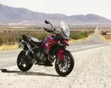 Czerwony motocykl Triumph Tiger 900 GT Pro zaparkowany na poboczu pustej drogi w krajobrazie z górami w tle.