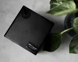 Czarny, nowoczesny dyfuzor zapachowy AROMA//PROJEKT z zielonymi liśćmi rośliny w tle.