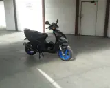 Czarny skuter Piaggio NRG MC3 z niebieskimi felgami stoi na betonowej posadzce w garażu. Obok niego leży zielony kask.
