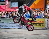 Motocykl do stuntu na tylnym kole, kierowca w czerwonej kurtce i kasku, tłum widzów na tle banerów.