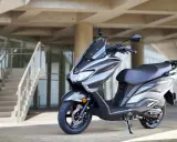 Szary skuter Suzuki Burgman, znany z tego, że to jedne z najbardziej bezawaryjne skutery, stoi przed nowoczesnym budynkiem z betonowymi schodami.