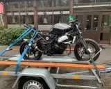 Czarny motocykl Kawasaki na przyczepce, zabezpieczony pasami. W tle budynek z napisem "Leśny Staw". Przepisy dotyczące przyczepki do motocykla są kluczowe dla bezpiecznego transportu.
