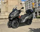 Trójkołowy skuter Peugeot Metropolis 400 na tle zabytkowej kamienicy i pomnika.