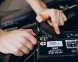 Ręce podłączają ładowarkę do akumulatora motocyklowego. Widać zacisk i wskaźniki na baterii.