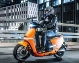Pomarańczowy skuter elektryczny z kierowcą w kasku i skórzanej kurtce jedzie nocą po miejskiej drodze.