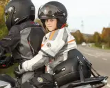 Uśmiechnięte dziecko na motocyklu, w kasku i kurtce, siedzi za kierowcą.