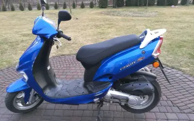 Błękitny skuter Kymco Vitality 50 2T stoi na brukowanym podjeździe, gotowy do jazdy.