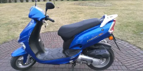 Błękitny skuter Kymco Vitality 50 2T stoi na brukowanym podjeździe, gotowy do jazdy.