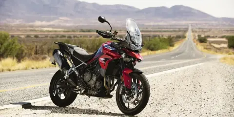 Czerwony motocykl Triumph Tiger 900 GT Pro zaparkowany na poboczu pustej drogi w krajobrazie z górami w tle.