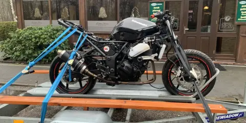Czarny motocykl Kawasaki na przyczepce, zabezpieczony pasami. W tle budynek z napisem "Leśny Staw". Przepisy dotyczące przyczepki do motocykla są kluczowe dla bezpiecznego transportu.