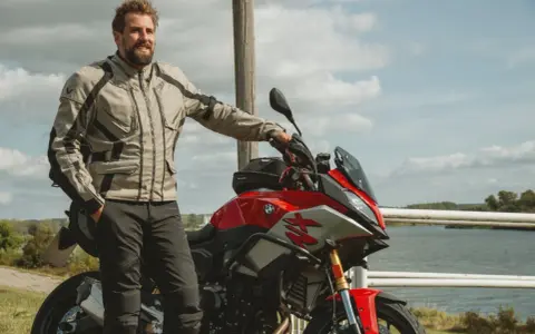 Uśmiechnięty motocyklista w kurtce od polskiego producenta odzieży motocyklowej, oparty o czerwoną maszynę nad jeziorem.