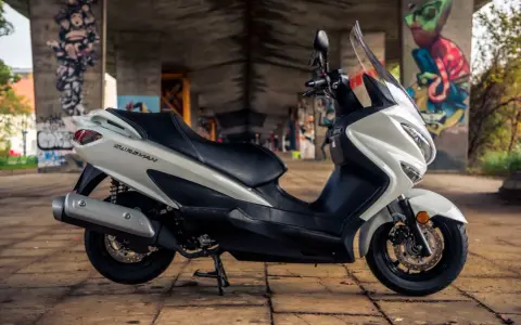 Biały skuter Suzuki Burgman 125, z czarnym siedzeniem, zaparkowany pod mostem z graffiti. Poznaj suzuki burgman 125 dane techniczne.
