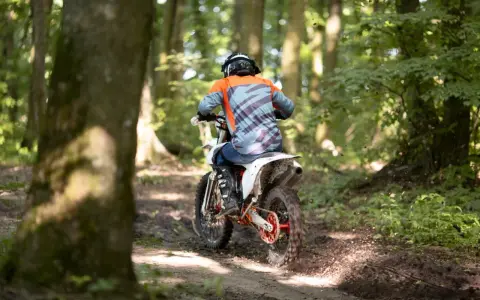 Motocyklista w kasku i pomarańczowej bluzie pędzi leśnym szlakiem. Od ilu lat można jeździć crossem? Odpowiedź zależy od przepisów i umiejętności.