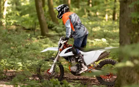 Motocyklista w kasku i pomarańczowej bluzie jedzie na białym crossie przez leśną ścieżkę.