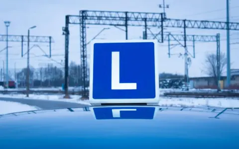 Tablica z literą "L" na dachu samochodu, symbolizująca naukę jazdy i teorię na prawo jazdy. W tle zimowy krajobraz z torami kolejowymi.
