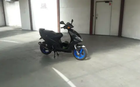 Czarny skuter Piaggio NRG MC3 z niebieskimi felgami stoi na betonowej posadzce w garażu. Obok niego leży zielony kask.