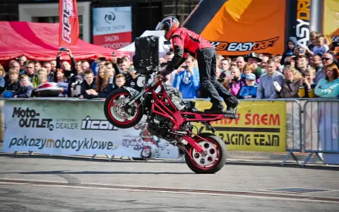 Motocykl do stuntu na tylnym kole, kierowca w czerwonej kurtce i kasku, tłum widzów na tle banerów.