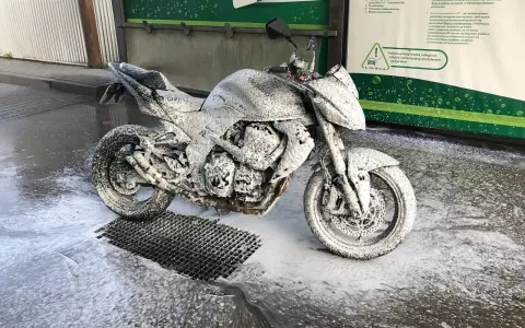 Motocykl pokryty pianą na myjni, gotowy do mycia. Instrukcja "jak myć motocykl" w tle.